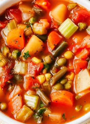 A bowl of hearty vegetable soup with fresh vegetables and herbs on a rustic table.